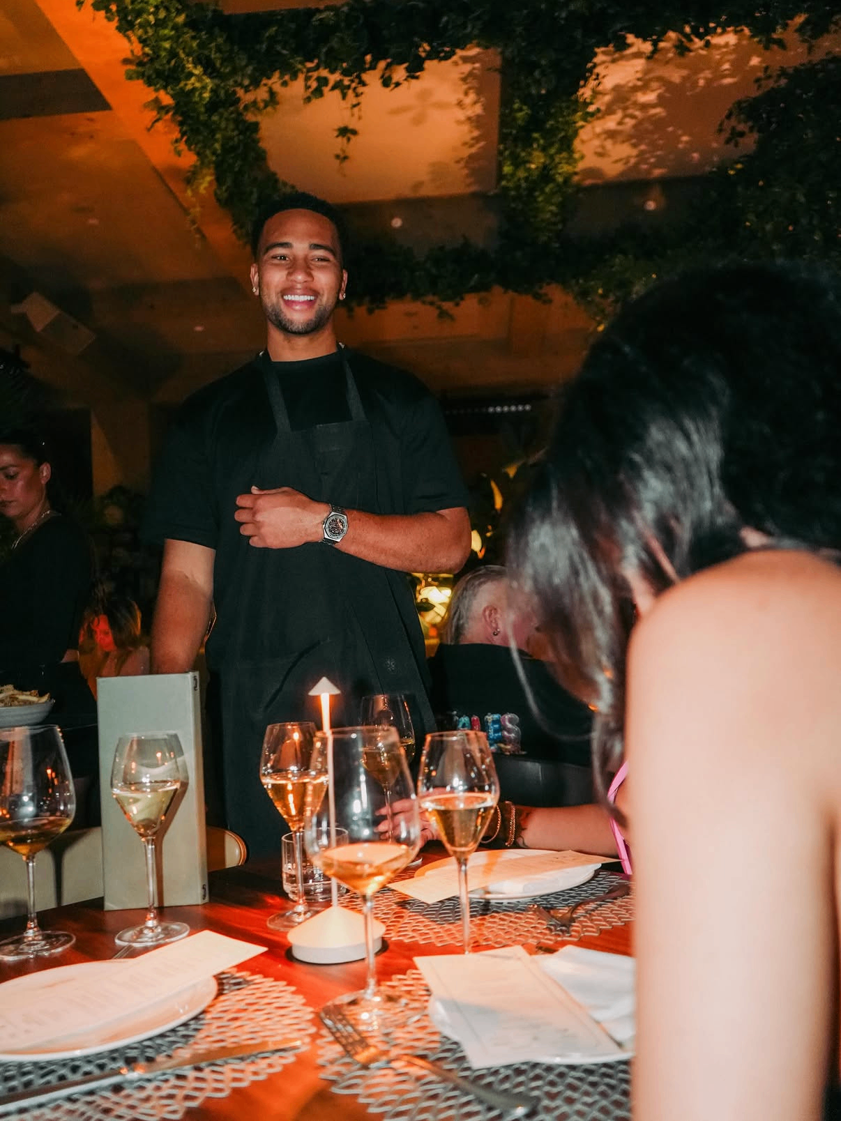 A celebrity waiter in a black apron smiles tableside while serving guests during the 2025 dinner service.