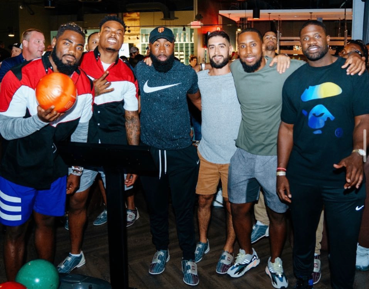 A group of celebrity players and guests pose together at the 2018 Bowl with the Pros Charity Event, with one player holding an orange bowling ball.