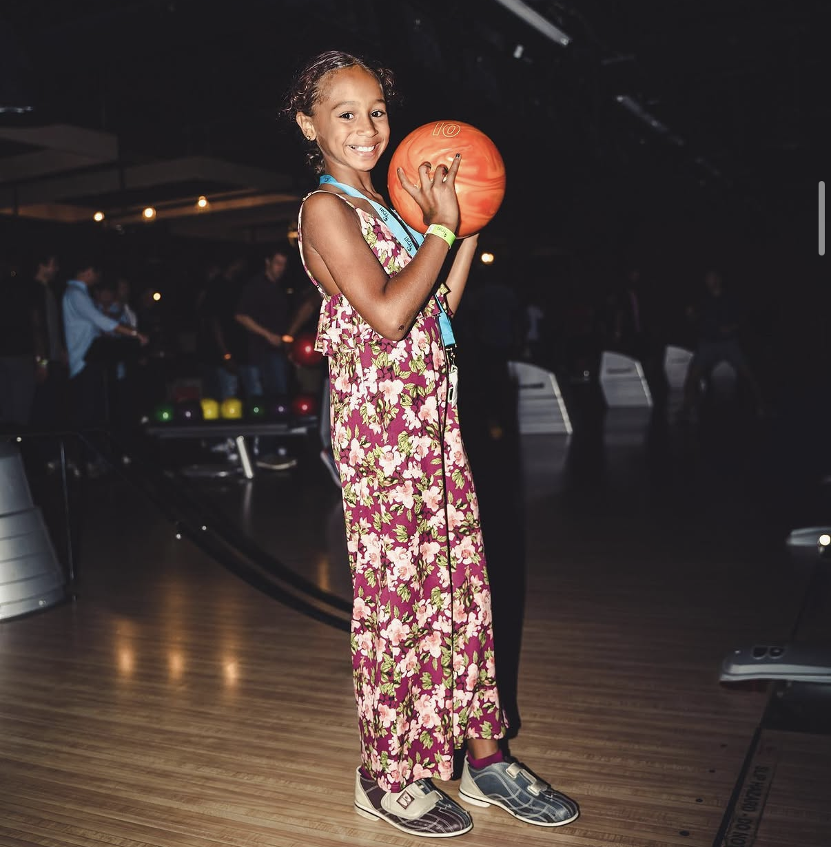 A young girl in a floral dress smiles and holds an orange bowling ball at the 2019 Bowl with the Pros Charity Event.