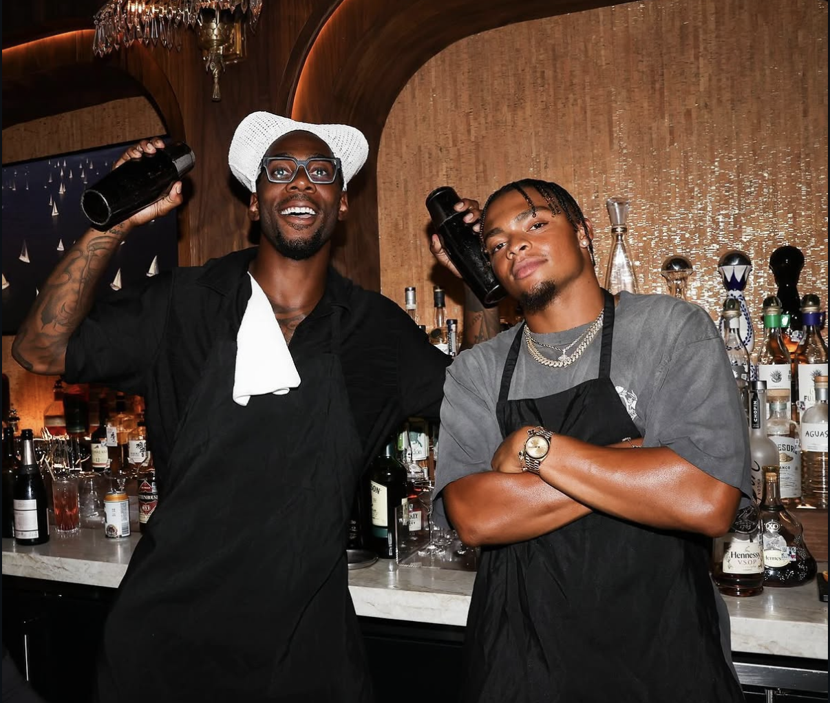Two celebrity bartenders pose behind the bar holding cocktail shakers.